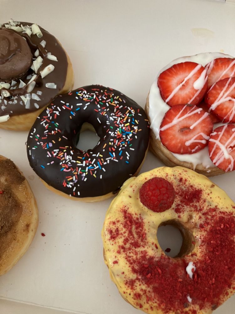 Fünf Donuts in einer weißen Pappschachtel. Sie liegen in Form einer Würfelfünf eng aneinander. Oben links Ein Donut mit Milchschokoladenglasur, dekoriert mit weißen Schokospänen und einem braunen Macaron oben auf.
Daneben einer mit weißer Schokoladenglasur, belegt mit fünf frischen Erdbeerscheiben und dünnen Zierstreifen aus hellem Guss.
Rechts unten liegt einer mit gelber Glasur, bestreut mit roten Himbeerbröseln und einer einzelnen, frischen Himbeere.
Unten links angeschnitten ist ein Donut mit Zimt-Zucker-Bestreuung.
Und in der Mitte ist ein Donut mit dunkler Schokoladenglasur platziert, gleichmäßig bedeckt mit bunten Zuckerstreuseln.