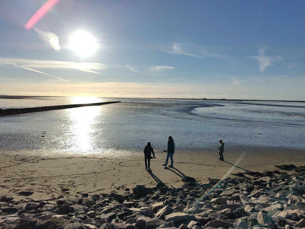 Drei Personen stehen an einem Nordseestrand. Das Wasser ist nicht da, es herrscht Ebbe. Der Himmel ist strahlend blau und die Sonne strahlt, so dass der nasse Schlick das Licht reflektiert.