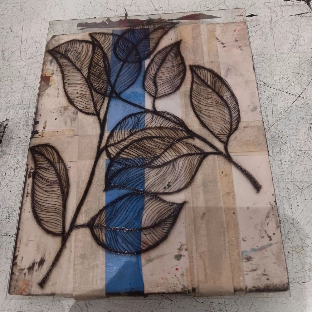 An inked up printing plate with a leaf design etched on. There are two leave twigs that overlap.