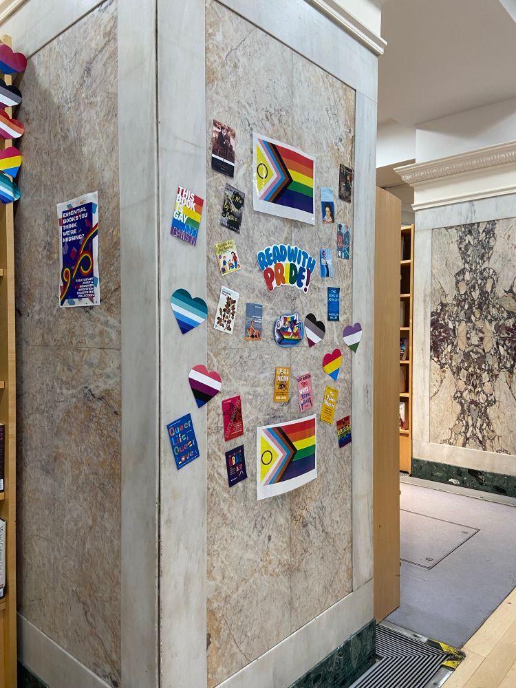 Awall display from a library with multiple heart-shaped Pride flags and posters reading “Queer Life, Queer Love” ￼and “Read With Pride”.