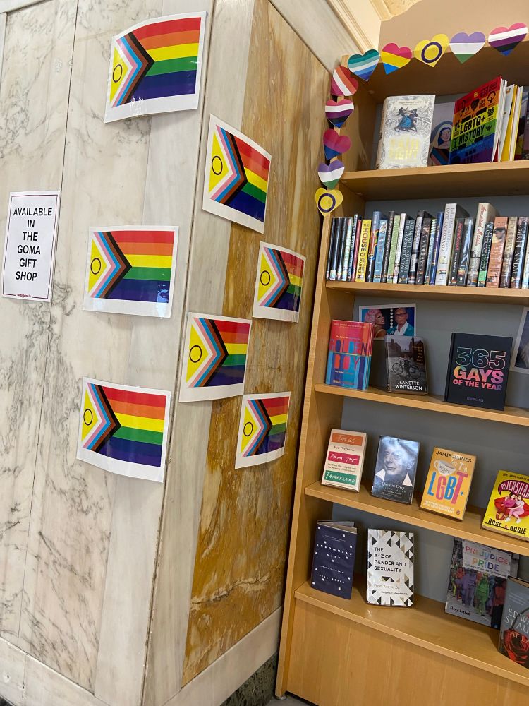 A display of library books with the rainbow Pride sticker on their spines and multiple Progress Pride flags stuck to the walls and shelves. A sign reads “Available in the GOMA gift shop”.