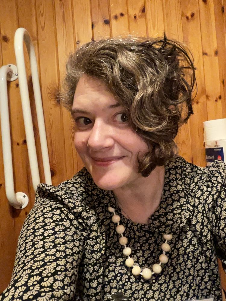 A selfie of a pale skinned enby wearing a 1950s style blouse and pearls with short dark curly hair 