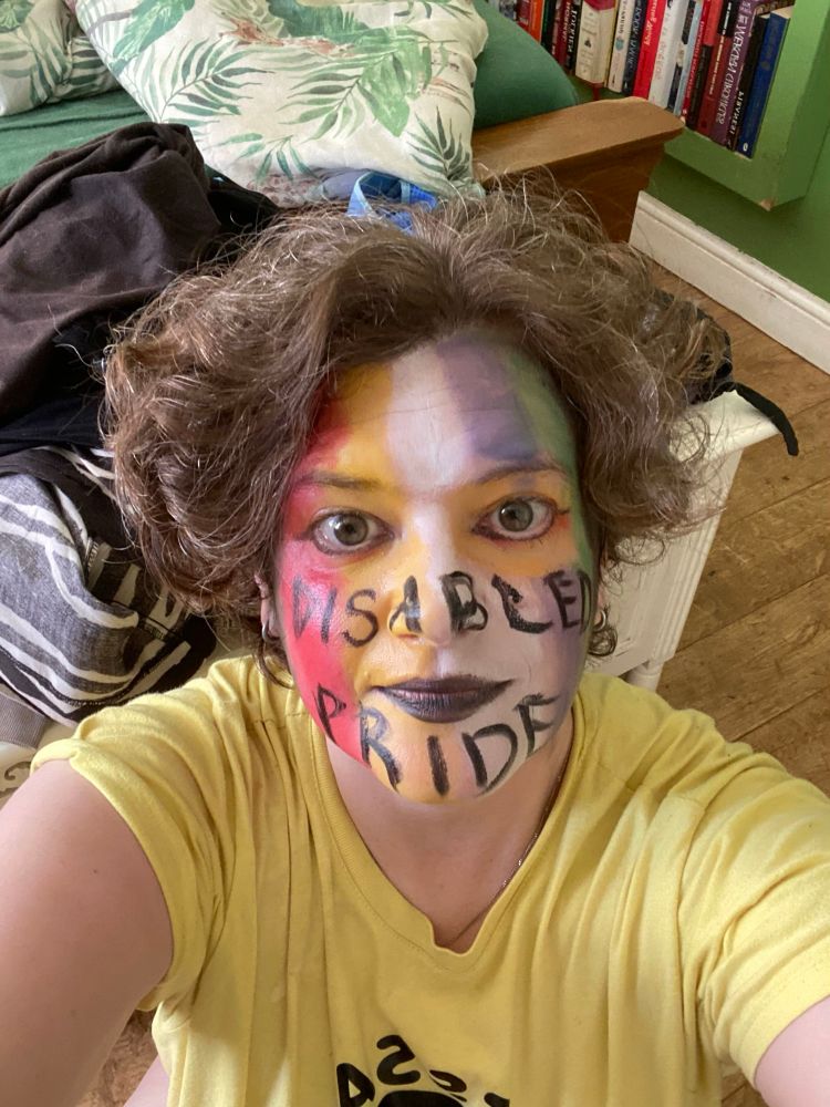 A pale-skinned enby with curly short auburn hair with their face painted in the colours of the Disabled Pride flag and the words “Disabled Pride” painted across their nose, cheeks and chin. They are wearing a yellow T-shirt. 