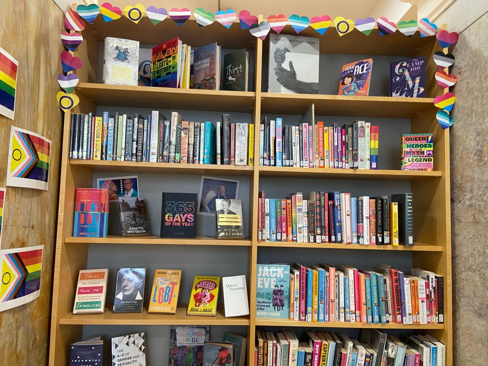 A display of library books with the rainbow Pride sticker on their spines. There are multiple Progress Pride flags stuck on the display shelves and walls and many other Pride identity flags shaped like hearts stuck on the shelves too.