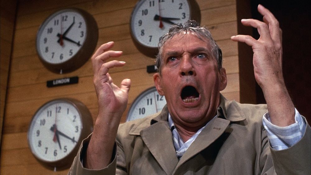 Peter Finch all wet from the rain holding his hands out like a mad prophet with clocks behind him in this still from Network