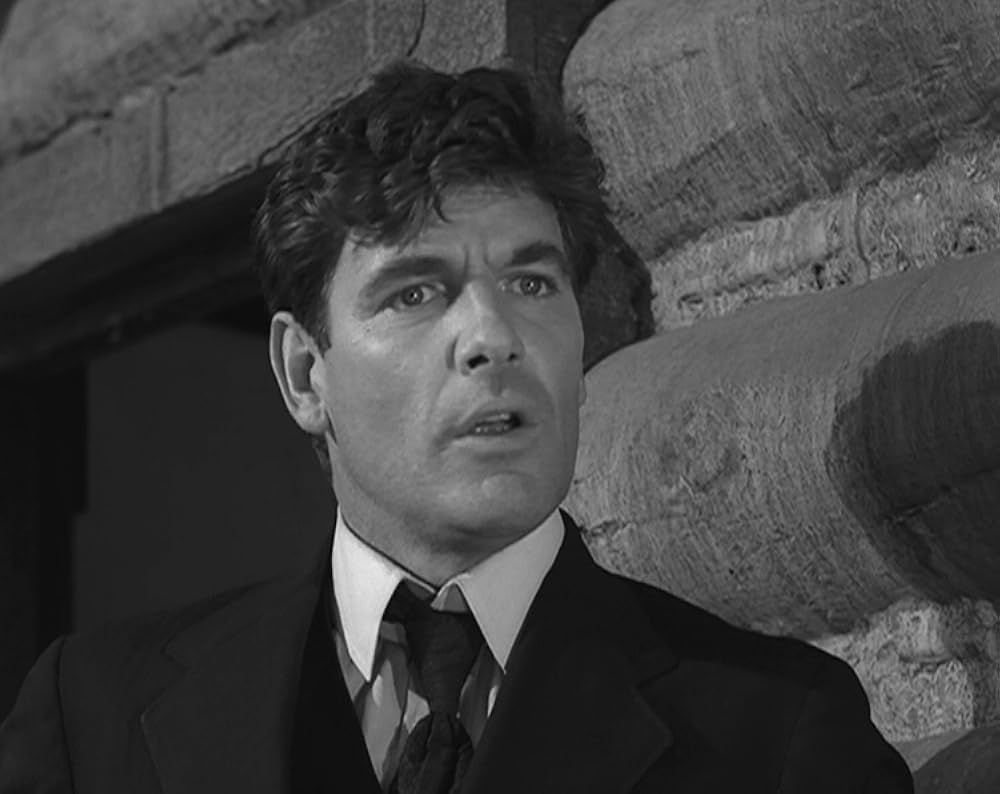 Black and white close up still of a white guy in a suit and tie with a log cabin behind him.