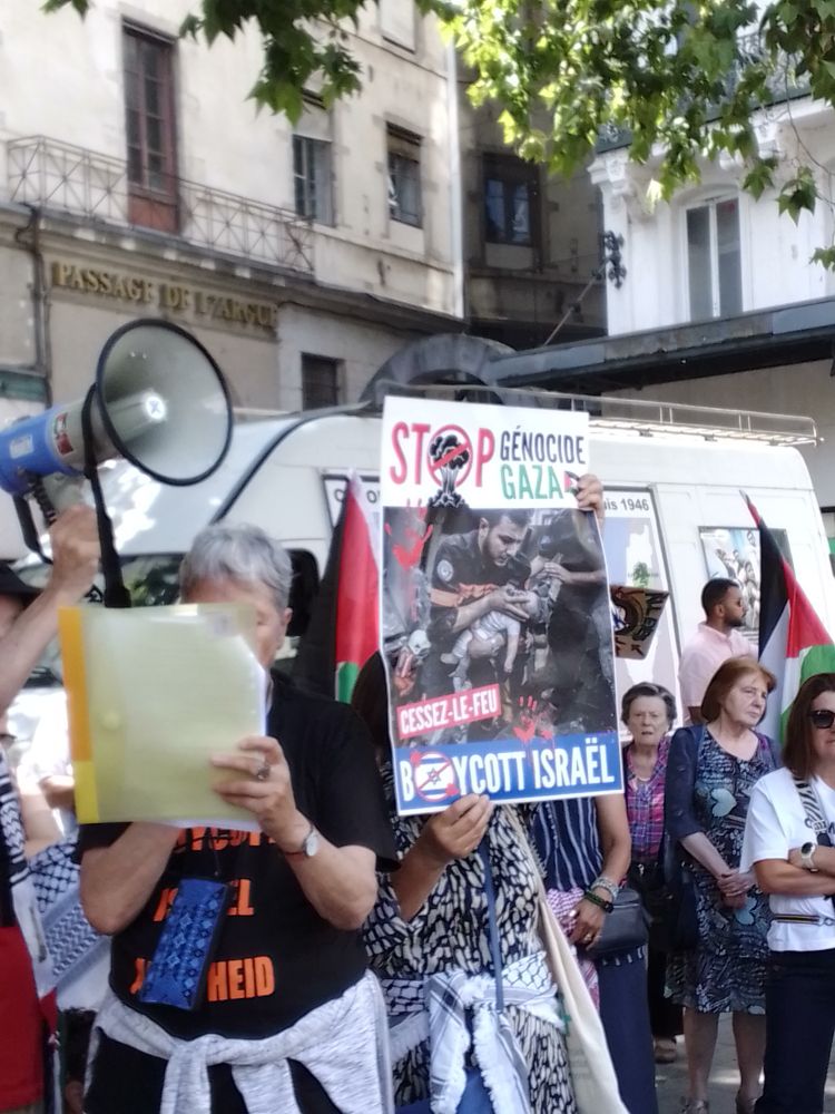 Prise de parole d'une militante de BDS (dont on ne voit pas le visage, caché par ses notes) avec à ses côtés une affiche réclamant le boycott d'Israël et exigeant un cessez-le-feu à Gaza tenue par une autre militante. 