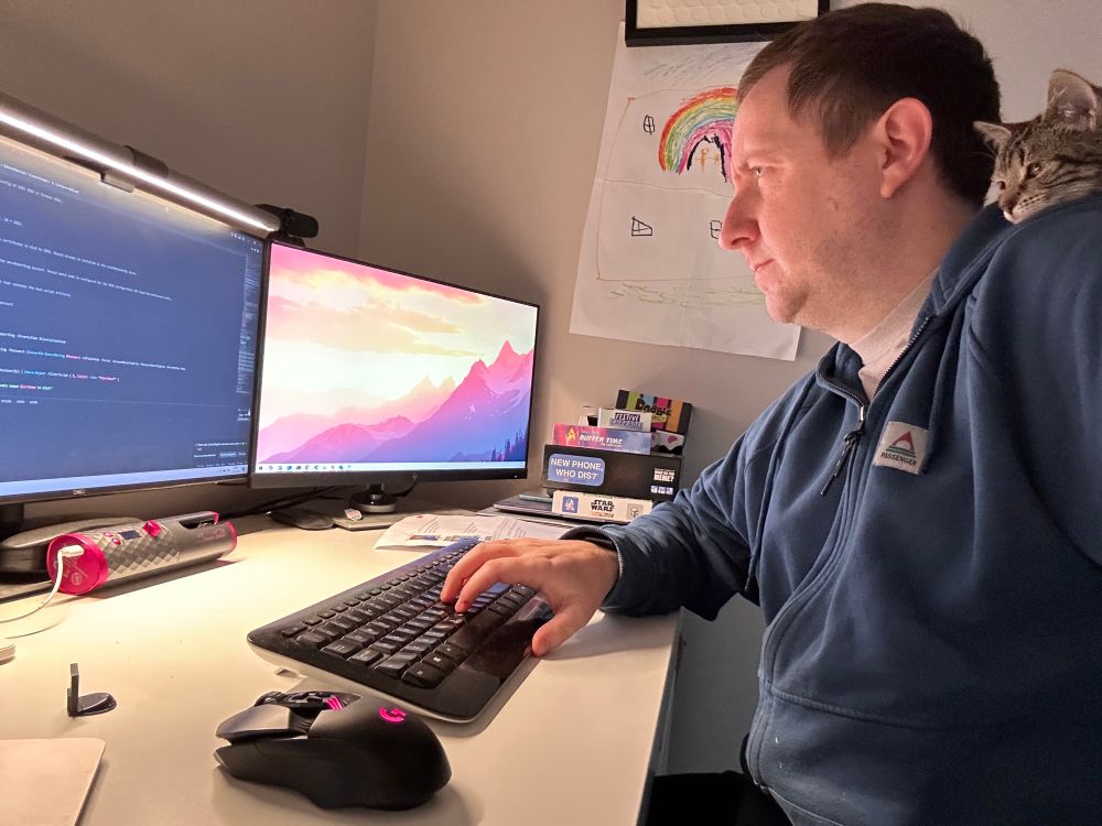A man sits as a computer working on code with a kitten on his shoulder