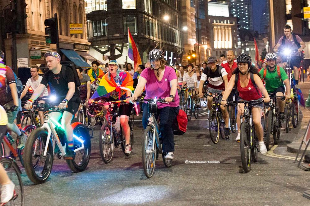 A group of people cycling together through the streets of London at dusk. People of all backgrounds some glad in brightly coloured capes and carrying Pride flags.