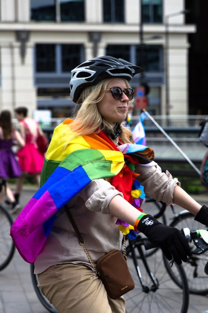 A smiling woman draped in a Pride flag riding a bicycle among a crowd of other cyclists with pride flags..