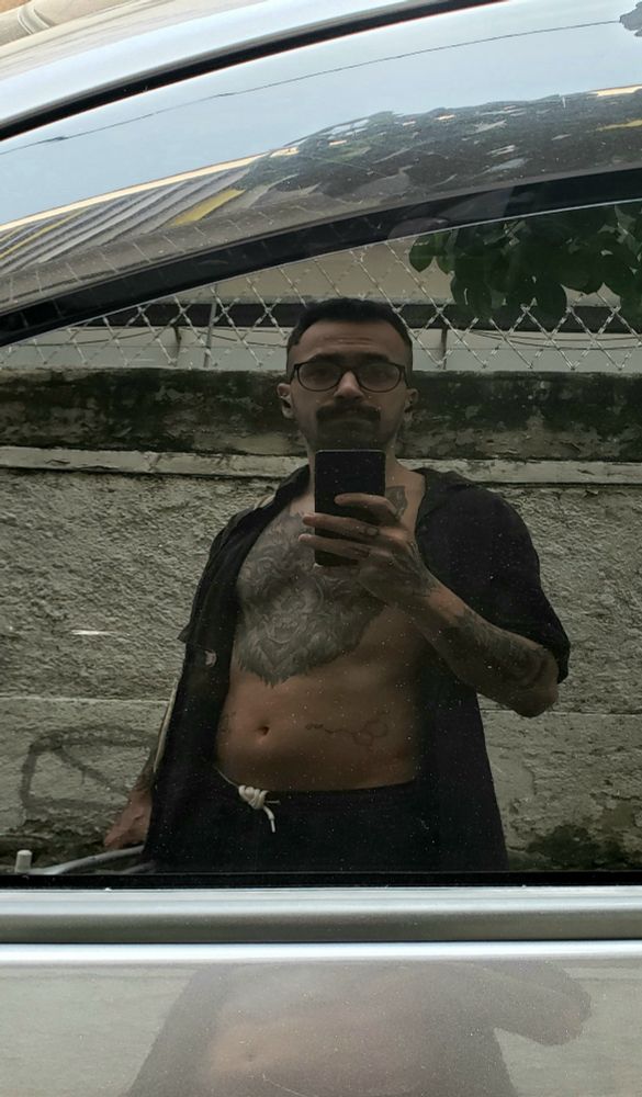 Foto de um homem tirada no reflexo de um carro. Ele tem várias tatuagens e segura uma cadeira de praia.
