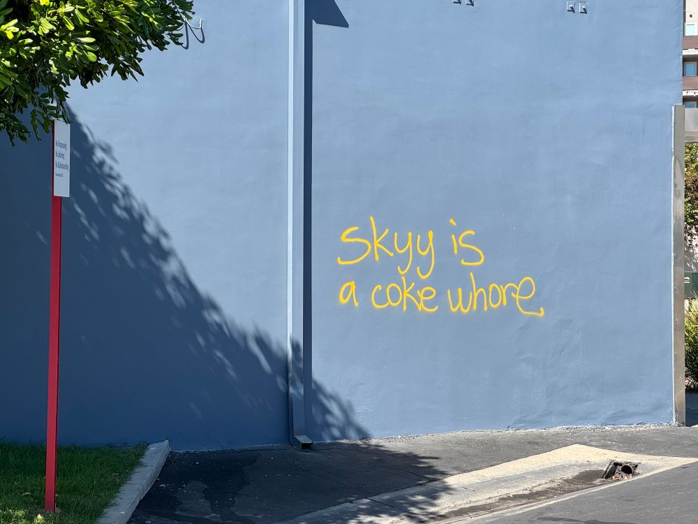 blue building outside wall with freehand spray paint graffiti in yellow, it says skyy is a coke whore. skyy has two y’s 