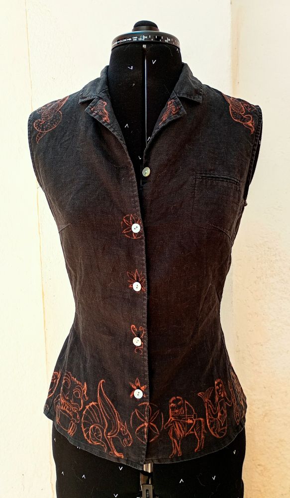 Fotografía de una blusa sin mangas con cuello de camisa en un maniquí vista de frente. Es negra con botones blancos y tiene dibujos en naranja óxido en la orilla inferior, alrededor de los botones y en los hombros.