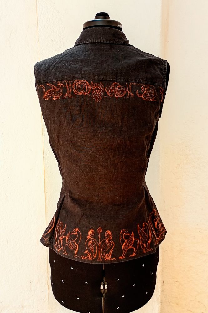 Fotografía de una blusa sin mangas con cuello de camisa en un maniquí vista de espalda. Es negra y tiene dibujos en naranja óxido en la orilla inferior y bajo la costura de la espalda.