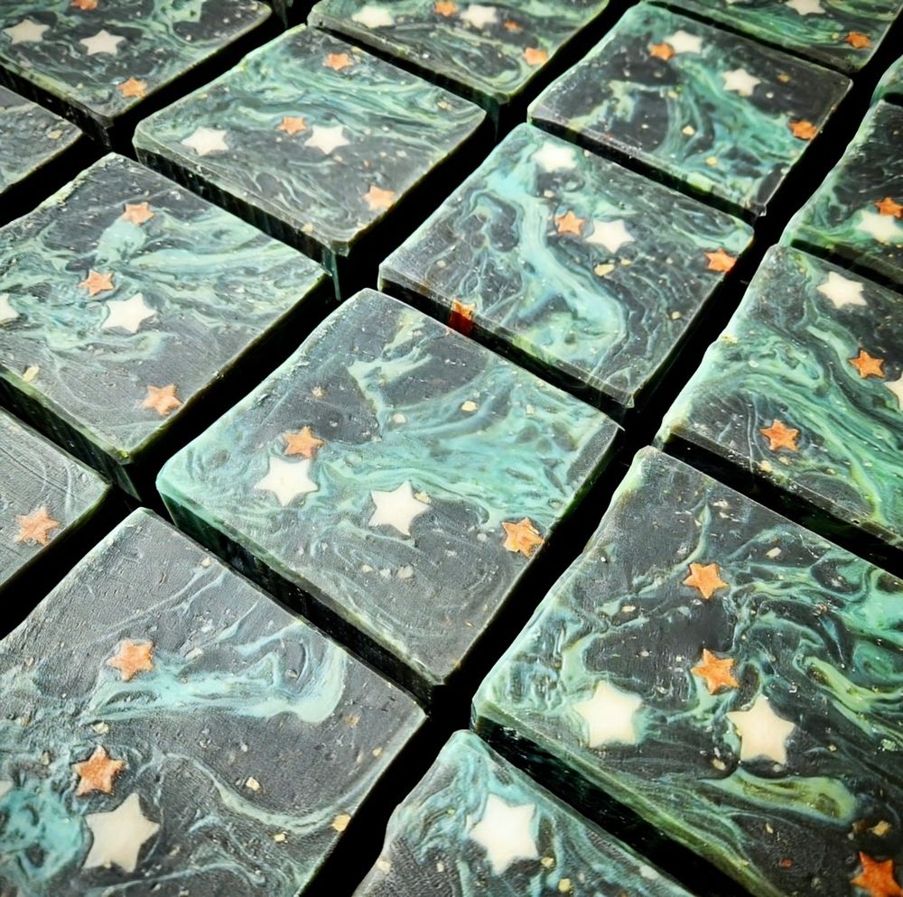 Handmade Soap in a nebula inspired design with a black background with swirls of blues and greens plus bits of colored asteroids, large white stars, and small orange/red stars. Featuring Cocoa Butter, Shea Butter, and Activated Charcoal with an alluring fragrance blend of bergamot, coconut milk, black pepper, cinnamon leaf, jasmine, tonka bean, cypress, cedarwood, and musk.