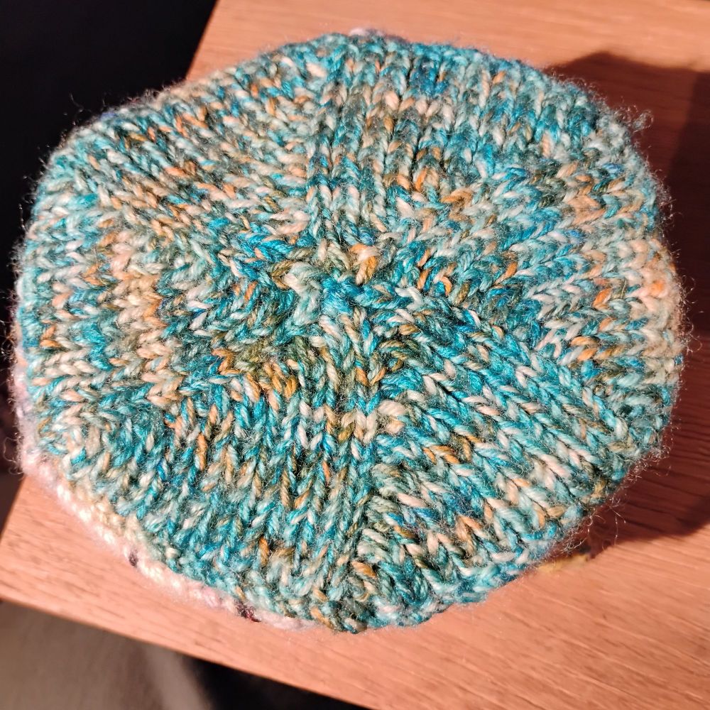 A knitted hexagon made from light turquoise yarn with orange speckles, on the corner of a wooden table.