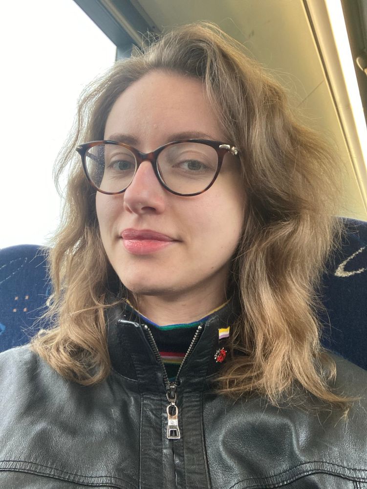 picture of me on a local link bus, wearing a black jacket, a ladybug pin and a nonbinary pride pin.