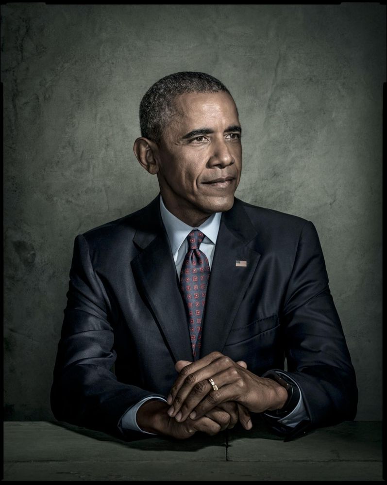 Portrait of President Barack Obama dressed in a classy suit and tie, and a US Flag pinned to his lapel.  Hands are crossed in front of him as he looks off a little to the left.