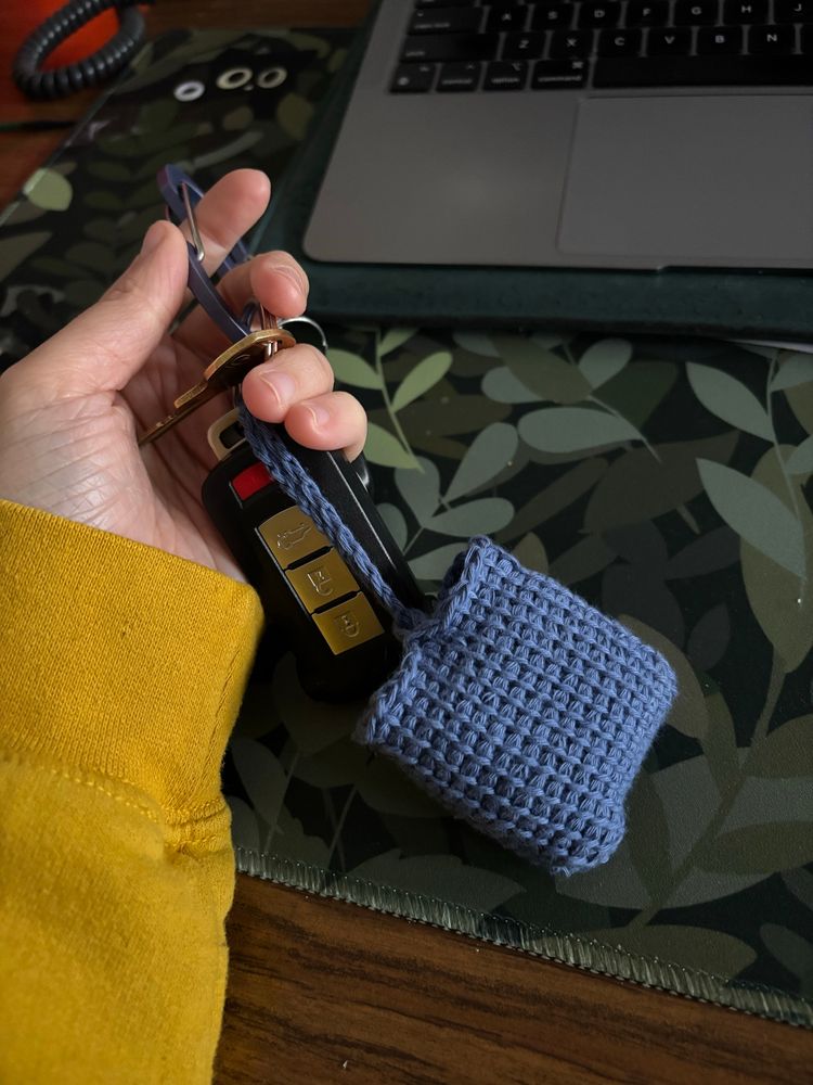 Hand holding keychain with blue crocheted AirPod case, yellow sweater visible on forearm 