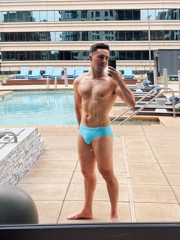 Selfie of a man (me) in a turquoise speedo taken in the reflection of a window in front of a pool
