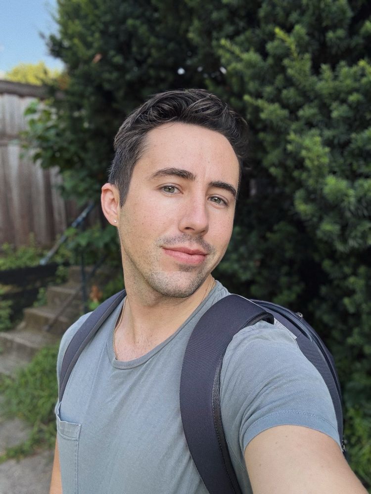 Selfie of a man (me) wearing a gray-green shirt and a black backpack, standing in front of an evergreen hedge 