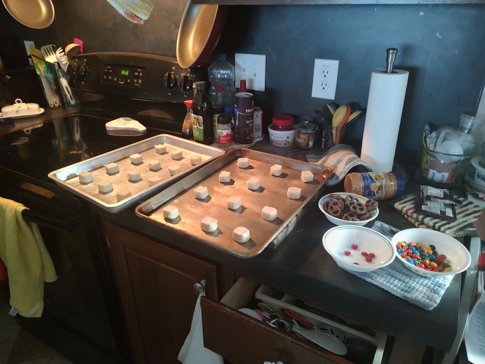 A cluttered kitchen counter and a black oven. The focus of the image is two cookie sheets covered with sugar cookie dough. Three bowls are full of chocolate covered pretzels and M&Ms