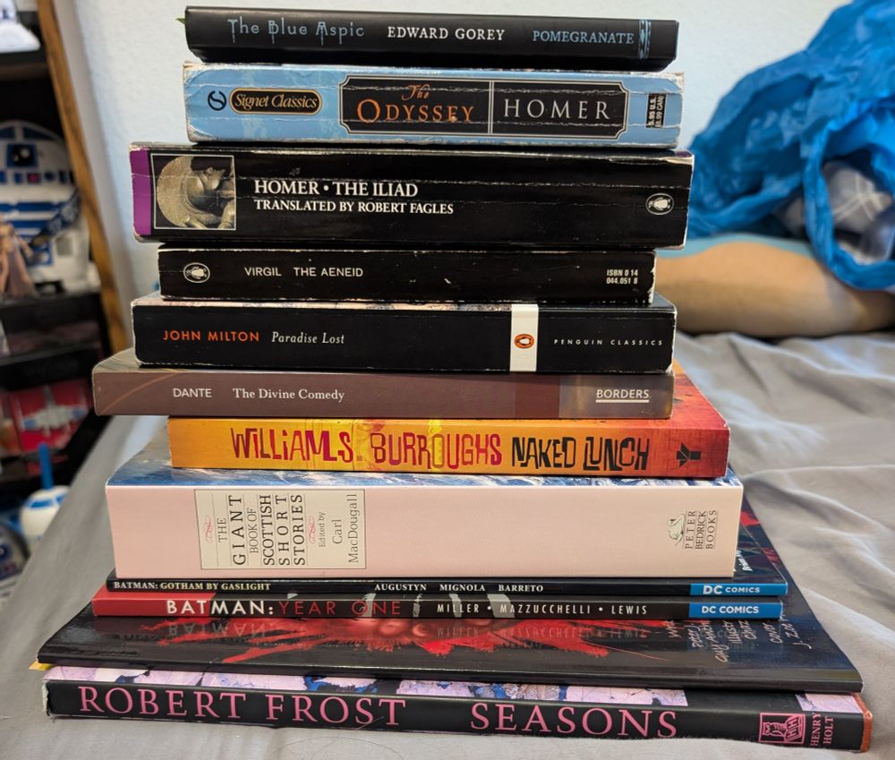 photo a a stack of books, from bottom to top: Seasons [Robert Frost], Behind These Eyes [Peter J Wacks & Anthony De Marco], Batman: Year One [Miller, Mazzucchelli, Lewis], Batman: Gotham by Gaslight [Augustyn, Mignola, Barreto], The Giant Book of Scottish Short Stories [edited by Carl MacDougall], Naked Lunch [William S. Burroughs], The Divine Comedy [Dante], Paradise Lost [John Milton], The Aeneid [Virgil], The Illiad [Homer], The Odyssey [Homer], The Blue Aspic [Edward Gorey]