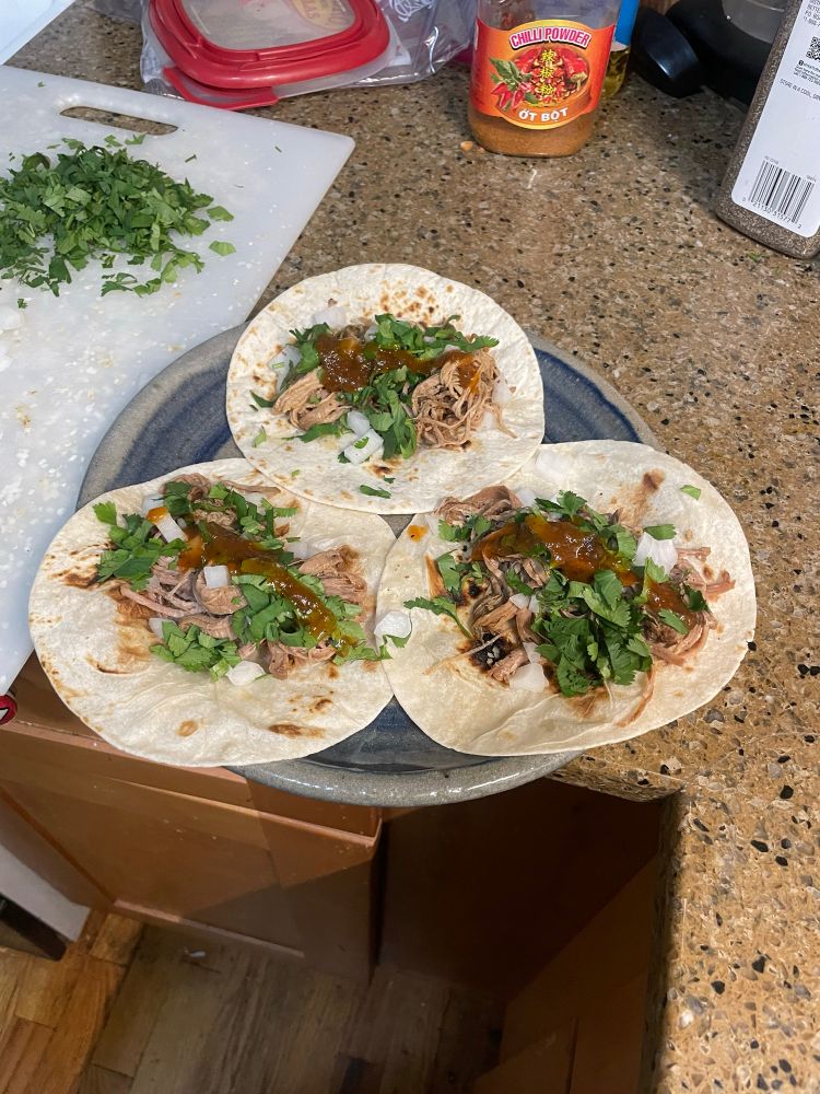 A plate of 3 shredded pork tacos, with onions, cilantro, and hot sauce