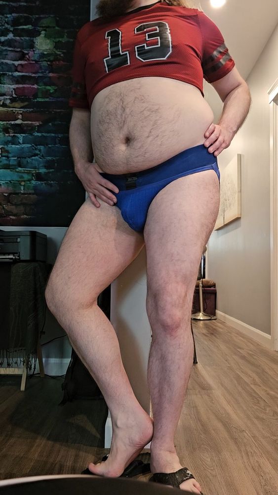 in a blue jockstrap and red sporty crob top, feet bare in flip flops