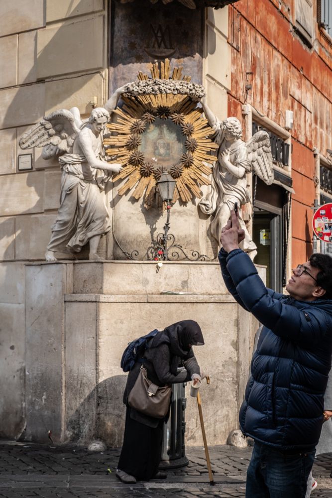 This photograph captures a moment on the streets of Rome, Italy featuring a religious shrine with two angel statues flanking a golden sunburst around an image. A man in a blue jacket is taking a picture of something in the distance, while a person, likely a beggar, dressed in dark clothing wearing a hood and leaning on a cane, stands in the background. The scene reflects a contrast between rich and poor, local and tourist, and tradition vs new technology.
