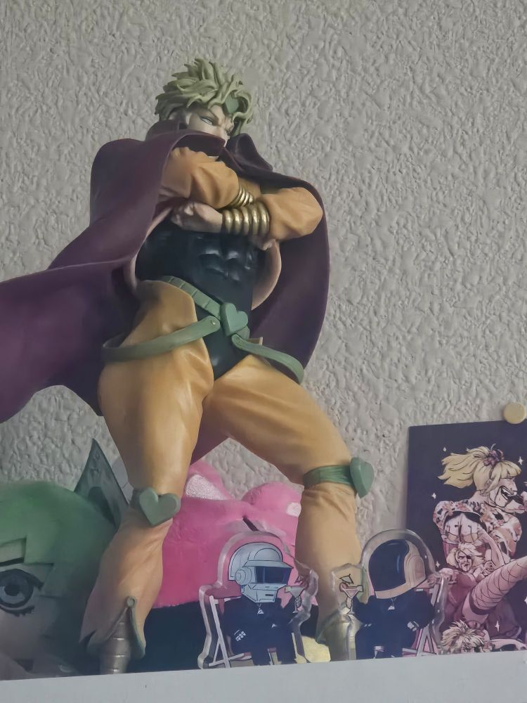 picture from below of a DIO statue. He has his maroon cape and is posing with his arms crossed and one hip popped. There are also some very small daft punk acrylic standees at DIO's feet