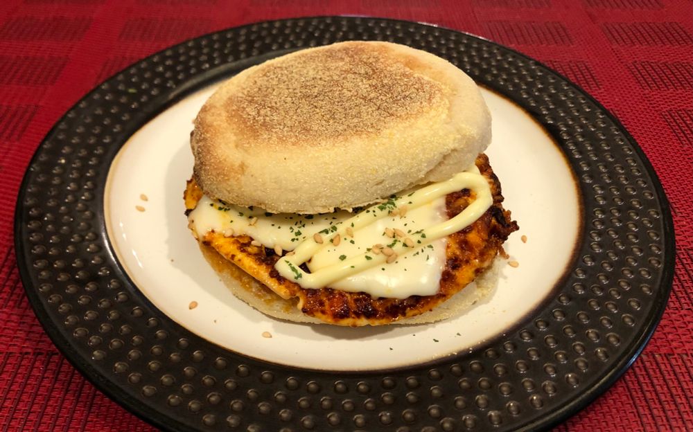 Photo of a chili crisp fried egg and cheese english muffin sandwich, on a brown and white plate, on a red placemat. The fried egg is dark orange/brownish in color from the chili crisp. Topped with a slice of melted white american cheese, some kewpie mayo, toasted sesame seeds and dried cilantro 