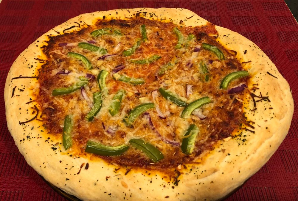 Photo of a round pizza on a plate, on a red placemat. The crust is pretty nicely browned, but sauce has escaped in one place just a bit where the crust dough wasn’t high enough. Toppings, that look kind of mixed in with the melted cheese, include sliced green pepper, red onion and garlic. 