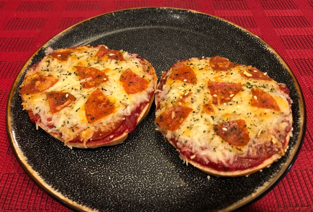 Photo of a pepperoni pizza bagel, on a black plate, on a red placemat. Homemade sesame bagel, cut in half. Topped with homemade pizza sauce, sliced pepperoni, mozzarella cheese, parmesan cheese, more pepperoni slices cut in quarters, dry pizza seasoning, dry parsley. Broiled in the oven until cheese is nicely browned/slightly burnt. 