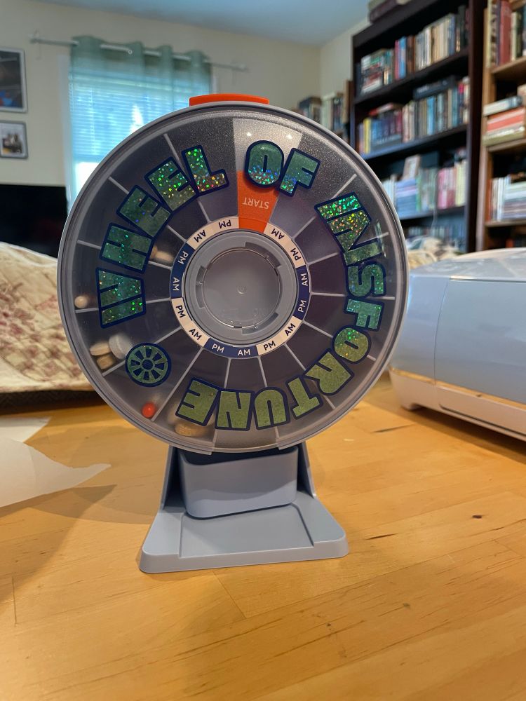 a circular push-button pill dispenser with a glittery circular sticker that says "wheel of misfortune" in the Wheel of Fortune font