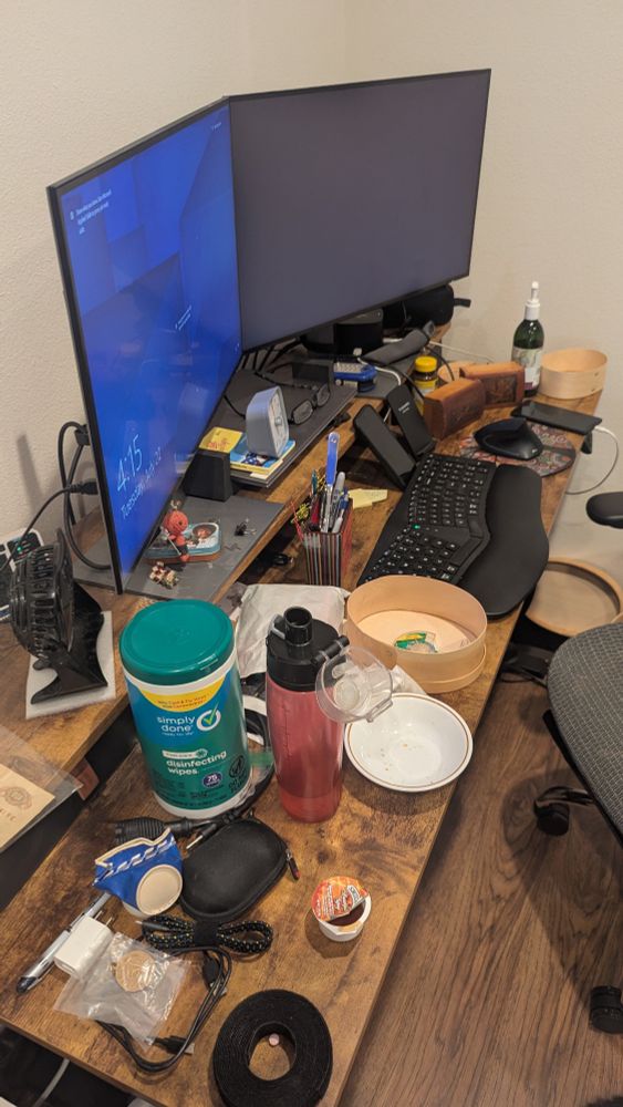 a long narrow desk with two computer monitors and a keyboard, entirely covered by various items, including empty food bowl, cleaning wipes, unidentifiable gadgets, small open decorative boxes, and who knows what else