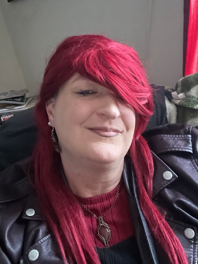 Alyssa is wearing her red hair side swept over her left eye to show off her guilloutine and skull earrings. She’s wearing a leather jacket and a maroon and black shirt. She has a labradorite pendant to center her look.