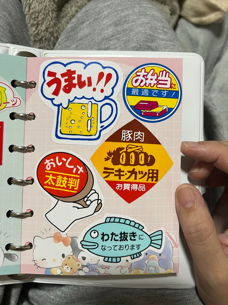 ポップな食品ラベル風シールのページ。「うまい!!」ビールジョッキ、「お弁当に最適です」、「豚肉 テキカツ用 お買得品」、「おいしさ太鼓判」、「わた抜きになっております（魚イラスト）」などが貼られている。背景にキティや小動物のキャラ。