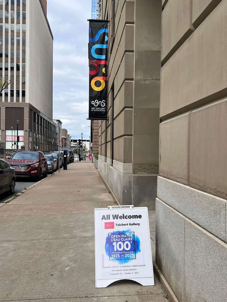 Shows the sidewalk sign for the Open Water 2025 Exhibition and the entrance of Teichert Gallery on Hollis street in Halifax.  The Art Gallery of Nova Scotia banner hangs on the wall in the middle distance. 