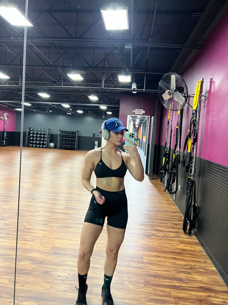Shelby posing in the gym mirror straight on in black shorts, a black sports bra, and a royal blue ATL hat. She is quite muscular 
