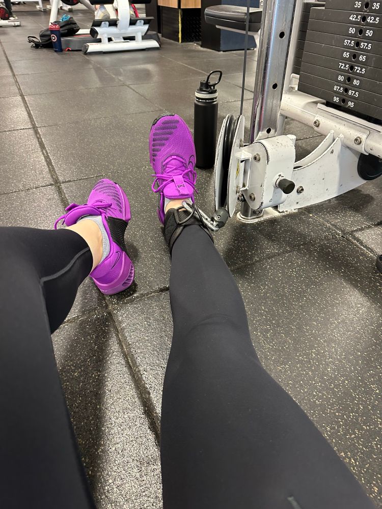 a pair of feet in tights and purple sneakers with the left leg wearing a large ankle strap that is attached to a gym weight cable