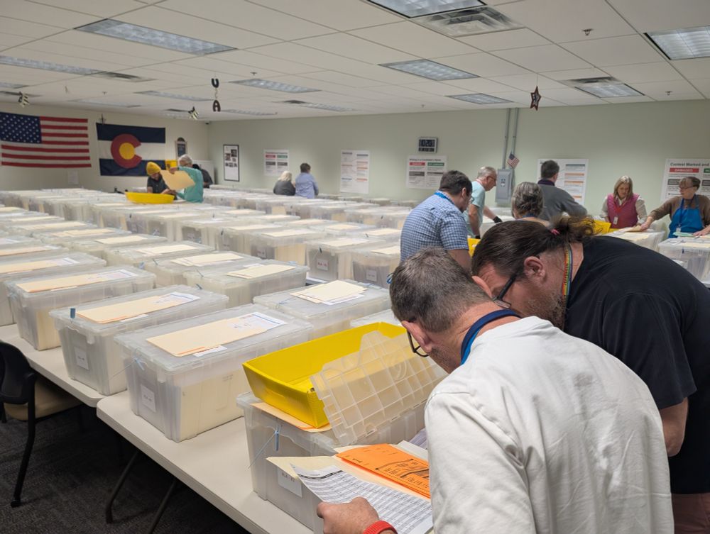 Two people lean over a folder with ballot scans and an open ballot bin. Dozens of bins on tables are in the background with other bipartisan pairs looking for specific ballots in them.