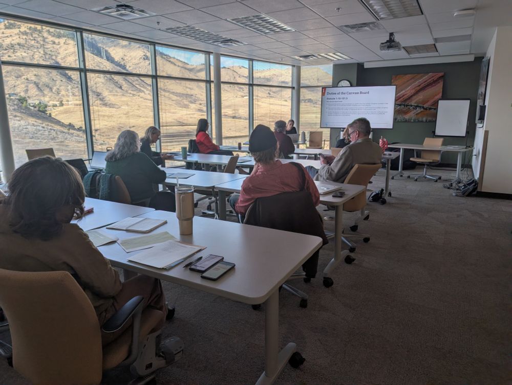 A group of people sit at tables facing a screen that reads "Duties of the Canvass Board" in a large-windowed conference room