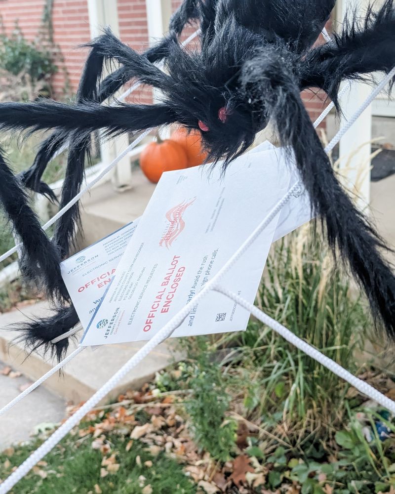 A large Halloween spider perches on a web made of twine holding two ballot envelopes with a house in the background