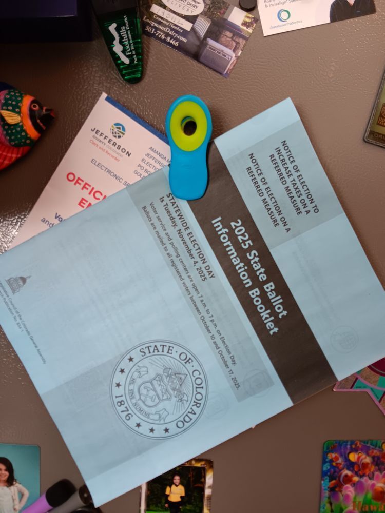 A Colorado blue book and Jeffco ballot envelope hang from a magnetic clip on a refrigerator surrounded by other magnets