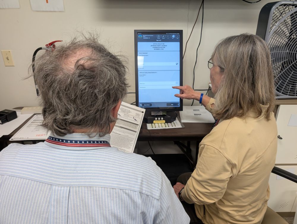 Two people sit at a screen with ballot questions shown on it. One person reads the paper ballot and the other makes selections on the screen.