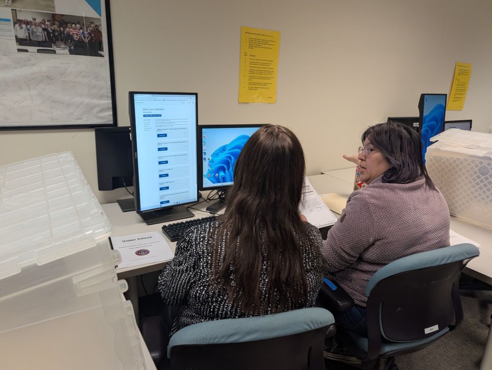 Two people sit at a computer and discuss a paper ballot they're holding with audit software on the screen in front of them