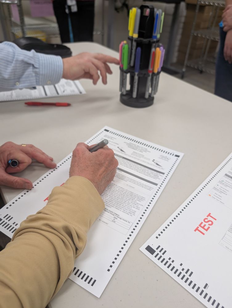 Someone's hands fill out a ballot with a colorful marker while another hand reaches for a new colored marker