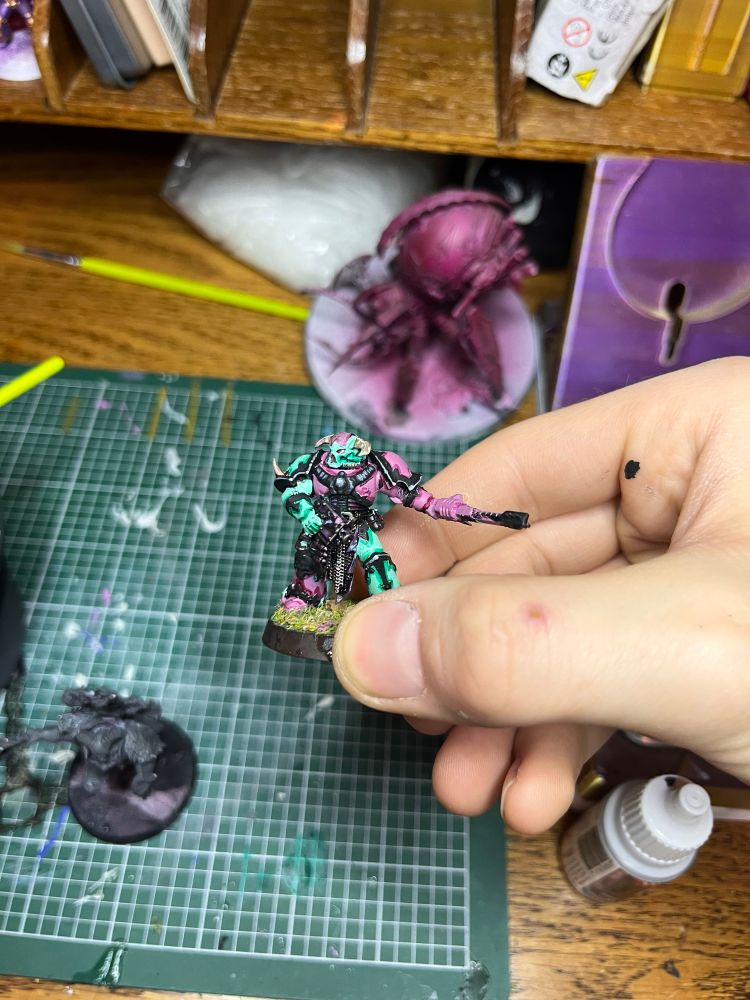 A warhammer miniature, specifically chaos space marine, holding a pistol and chainsword. It’s painted teal, pink, and black, and held above a desk. 