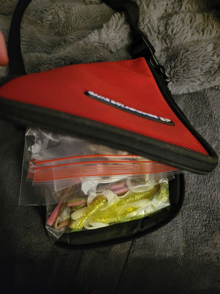 The long, rectangular, red bag has been opened and inside there are ziploc bags with fishing baits and lures inside of them. There are glittery yellow and pink rubber pieces.